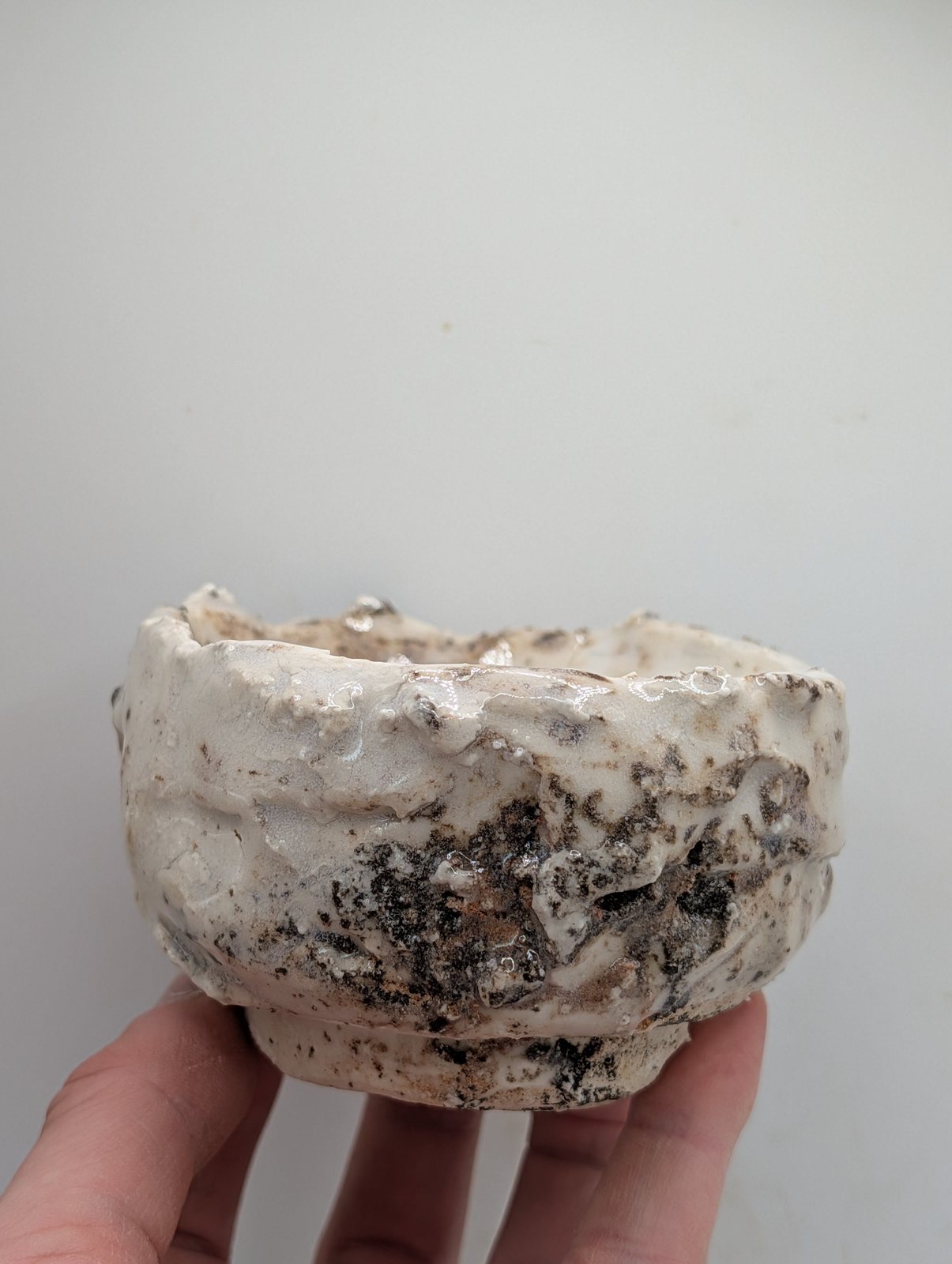 A small handbuilt cup with a white crackled glaze and dark speckling, held in a hand against a plain wall.